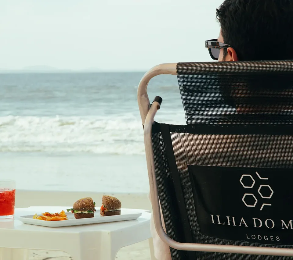 Cadeira com a logo da pousada na areia, com drinks e petiscos numa mesa diante do mar