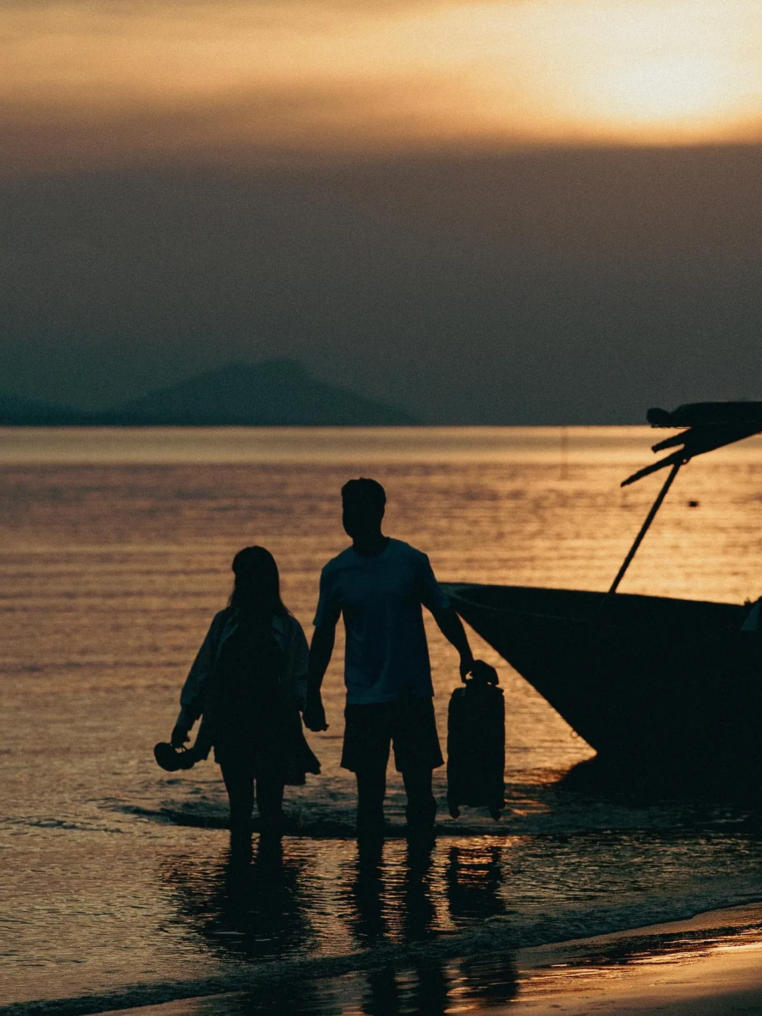 Casal caminha na água rasa ao pôr do sol, com a mala na mão, ao lado do barco que os trouxe