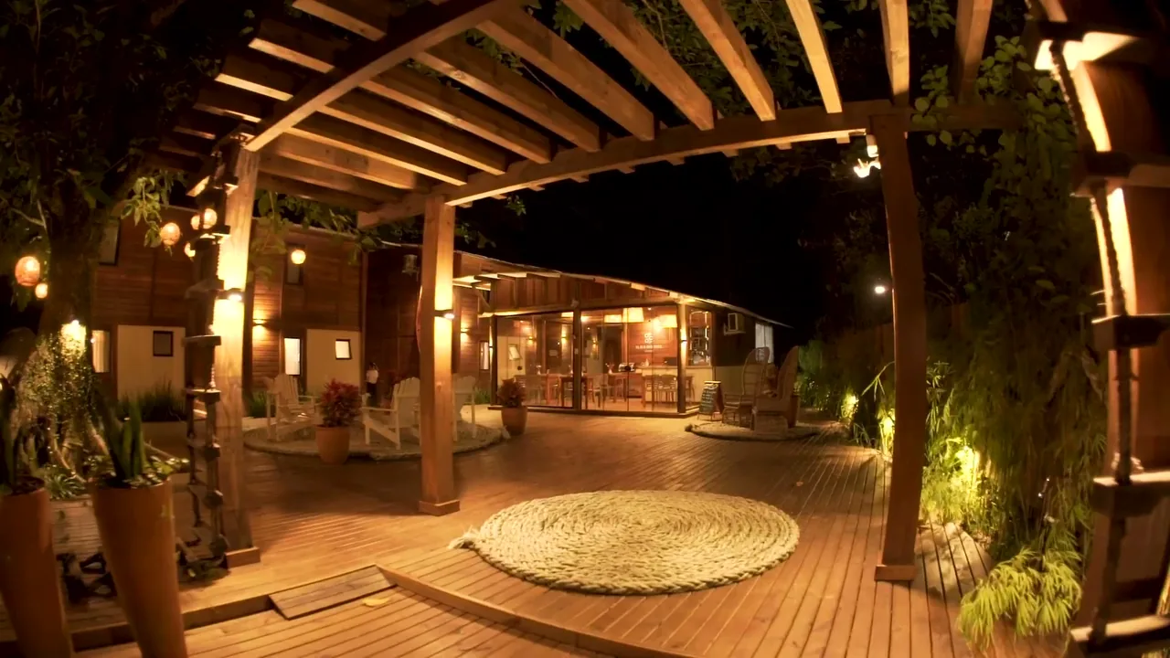 Deck de madeira iluminado à noite, com tapete de corda, poltronas e o bistrô ao fundo