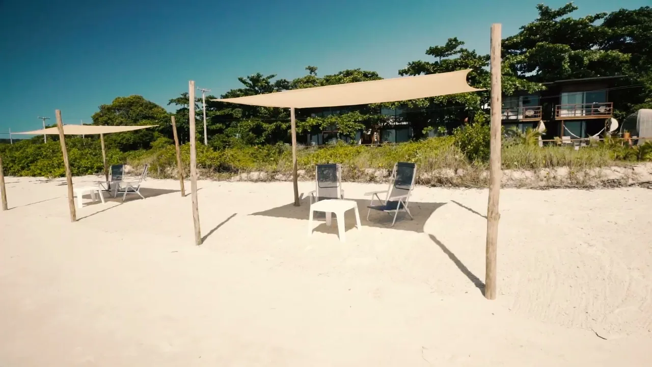 Toldos de lona e cadeiras na areia branca, com os lodges entre as árvores ao fundo